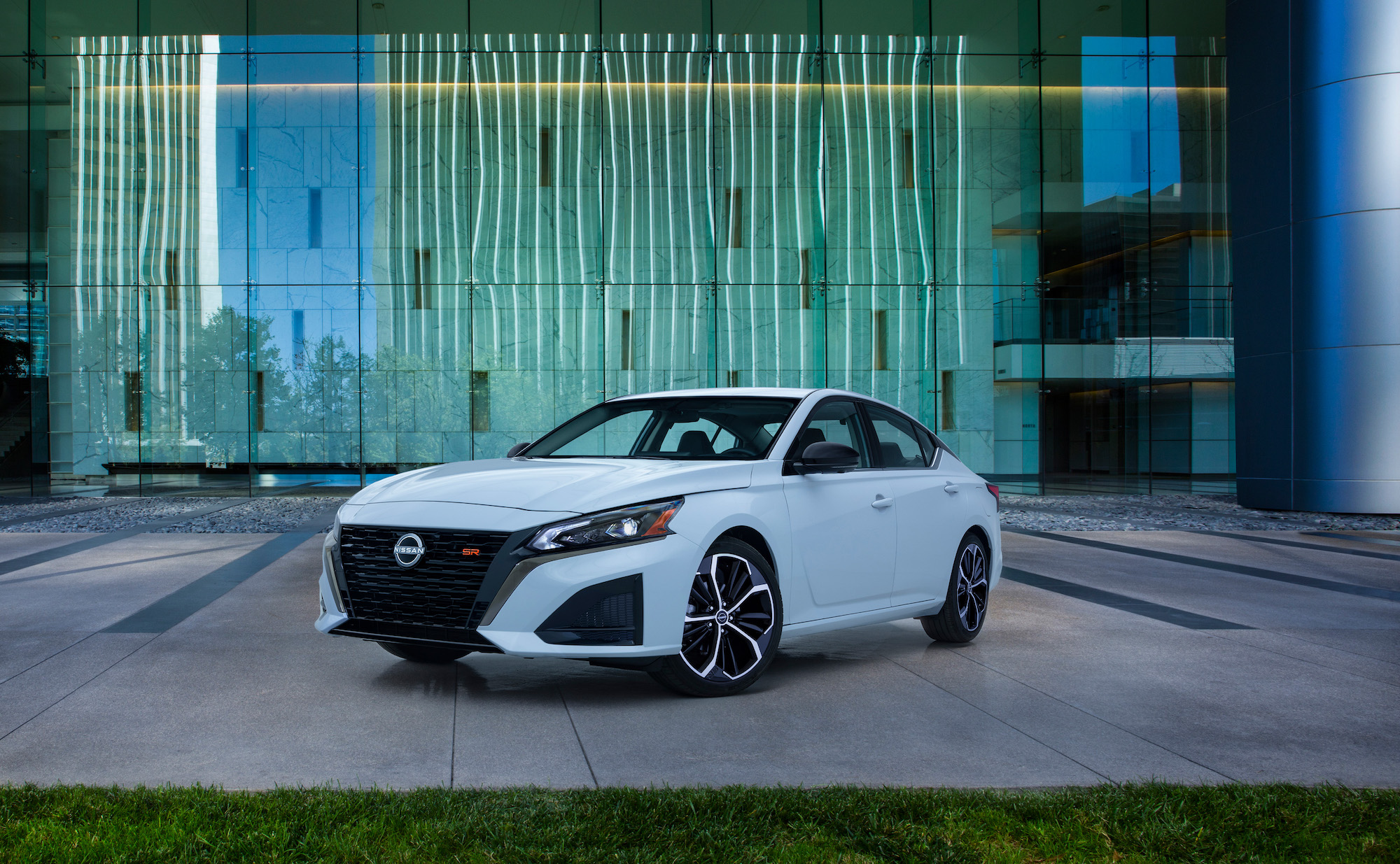 A white 2023 Nissan Altima, a good family car, sitting in front of a green lined wall on pavement.