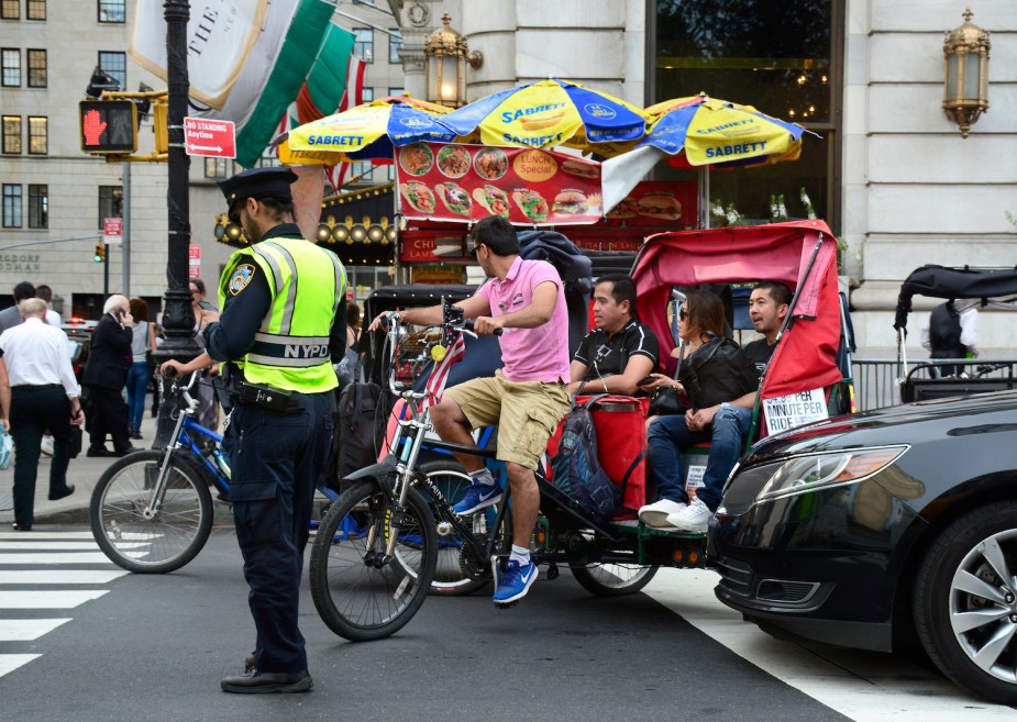 Bicycles, rickshaws, and cars waiting for a light to change at a busy intersection, a NYPD police officer visible in the foreground.