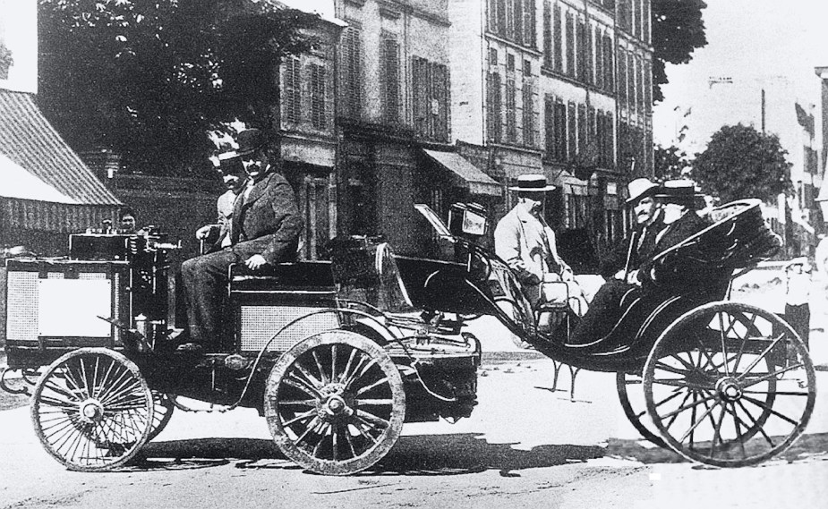 Picture of a steam-powered "tractor" with trailer parked in Paris in 1894, buildings and a city street visible in the background.