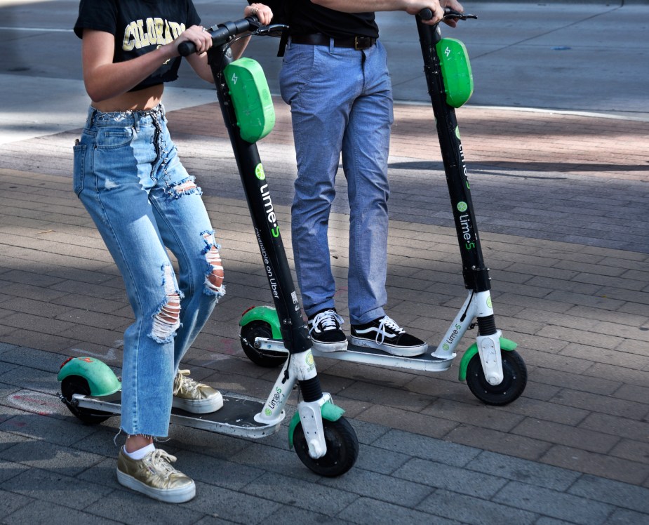 A closeup of two Lime-S kick scooters and the feet of their riders, a cobblestone street visible in the background.