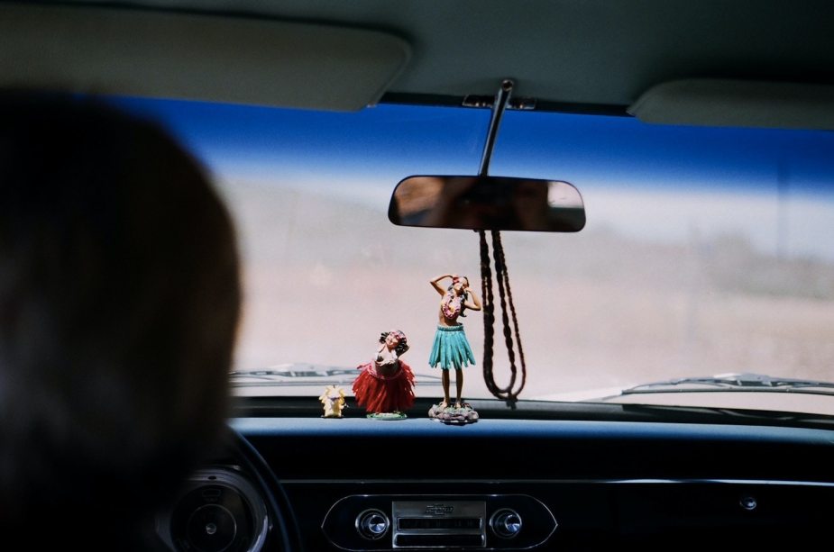 Image of two hula girl figurines on an old cars dashboard, the road beyond the windshield is out of focus and blurry.