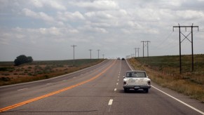 An old white station wagon car driving down an empty rural road with telephone poles and fields visible in the background.