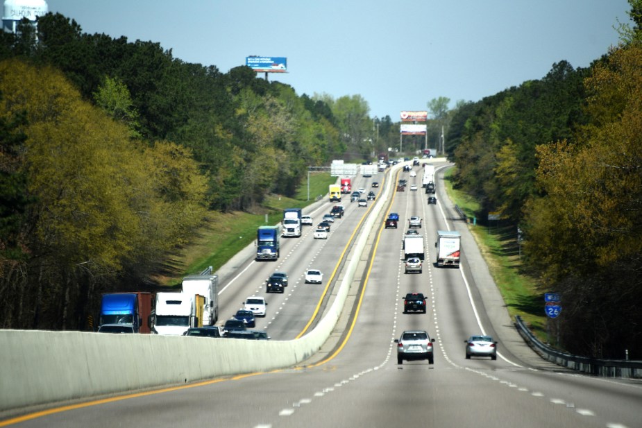 Drivers in the left lane of a busy interstate highway lined by trees and billboards.