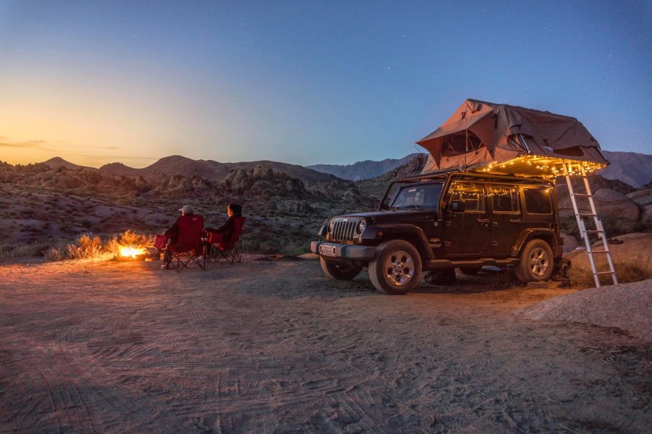 Black four-door Jeep Wrangler parked in the desert by a campfire with a rooftop tent above it, campers sitting nearby.