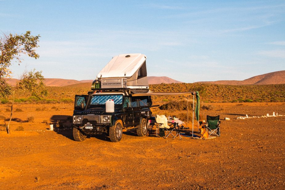 Black land rover Defender parked in a sandy campsite with a rooftop tent popped, a ridge of mountains visible in the background.