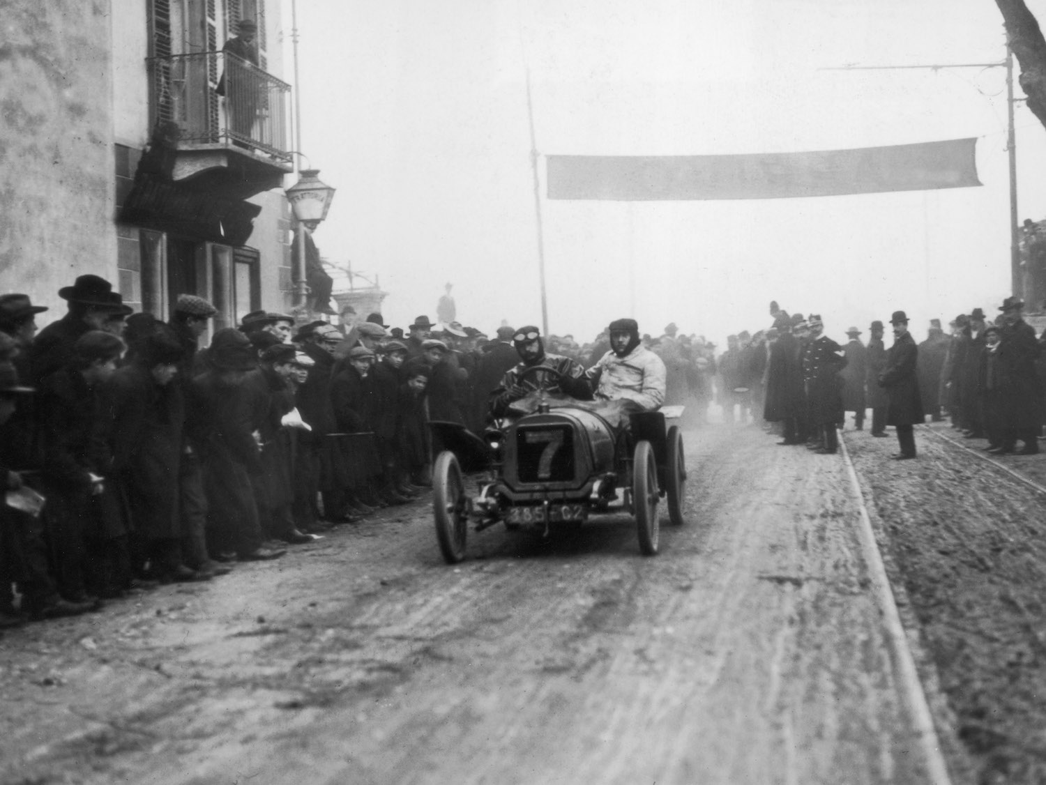 Two men finishing a race in an early Peugeot grand prix car on a muddy track, the finish line crowd visible in the background.