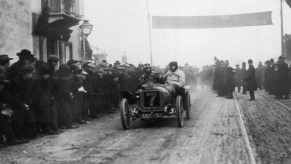 Two men finishing a race in an early Peugeot grand prix car on a muddy track, the finish line crowd visible in the background.