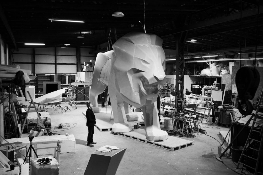 A twenty-foot-tall lion sculpture still in a workshop with an artist staring up at it.