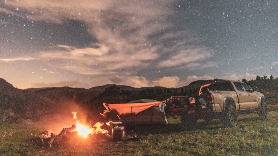 Shot of a Tacoma pickup truck parked in the mountains, a tent set up next to it and a bonfire behind its tailgate.