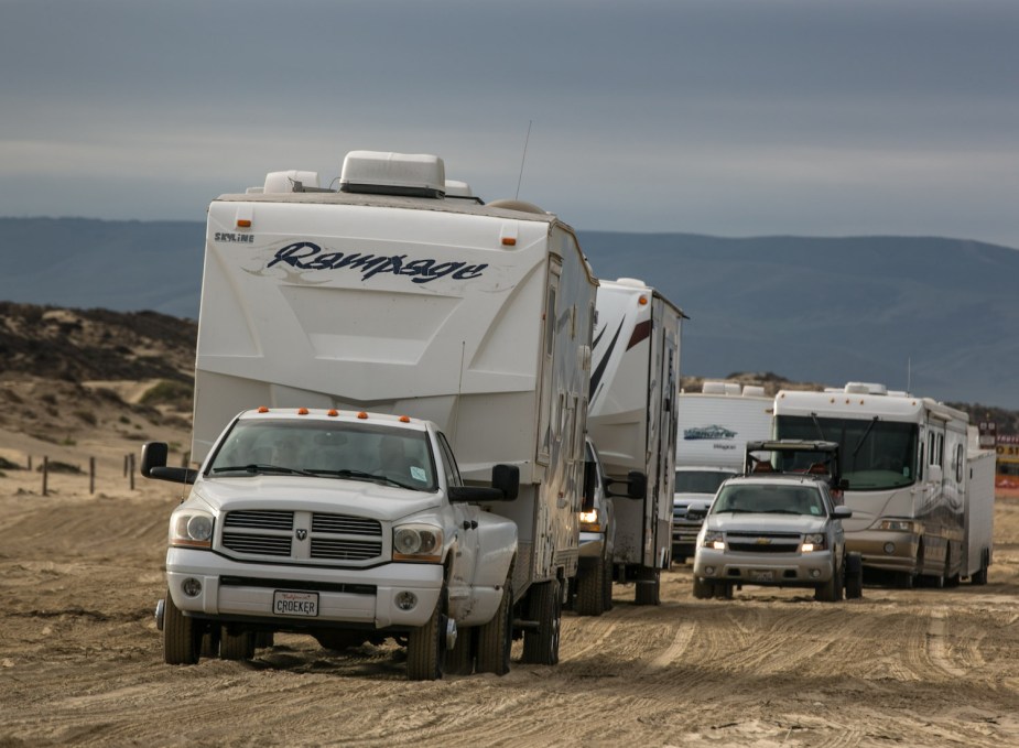 A convoy of trucks and SUVs tow campers onto Pismo Beach in California, mountains visible in the background.