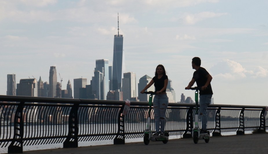 A couple rides their Lime electric kick scooters along a shoreline, the new york city skyline visible in the background.