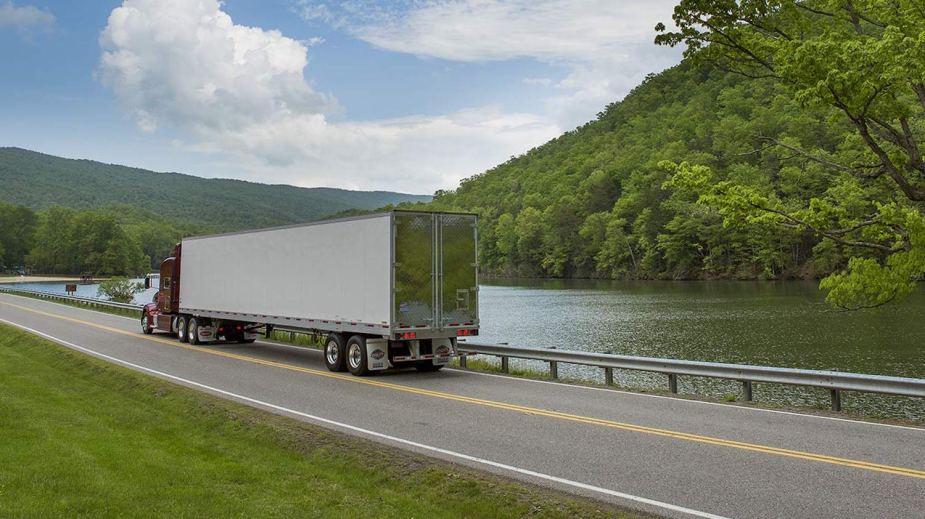 A promo photo of a semi truck and trailer driving along a rural road, a river and mountains visible in the background.