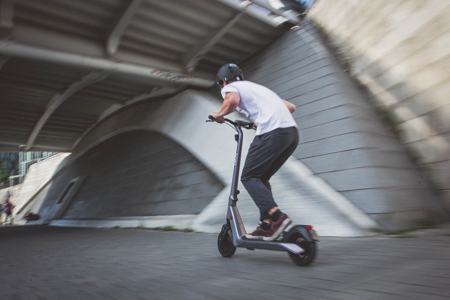 A man in a helmet races under a bridge on an electric kick scooter, a blurry stone wall visible in the background.