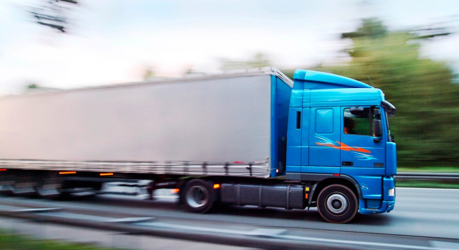 A blue semi truck tractor hauling a white trailer down a German highway, blurred trees visible in the background.