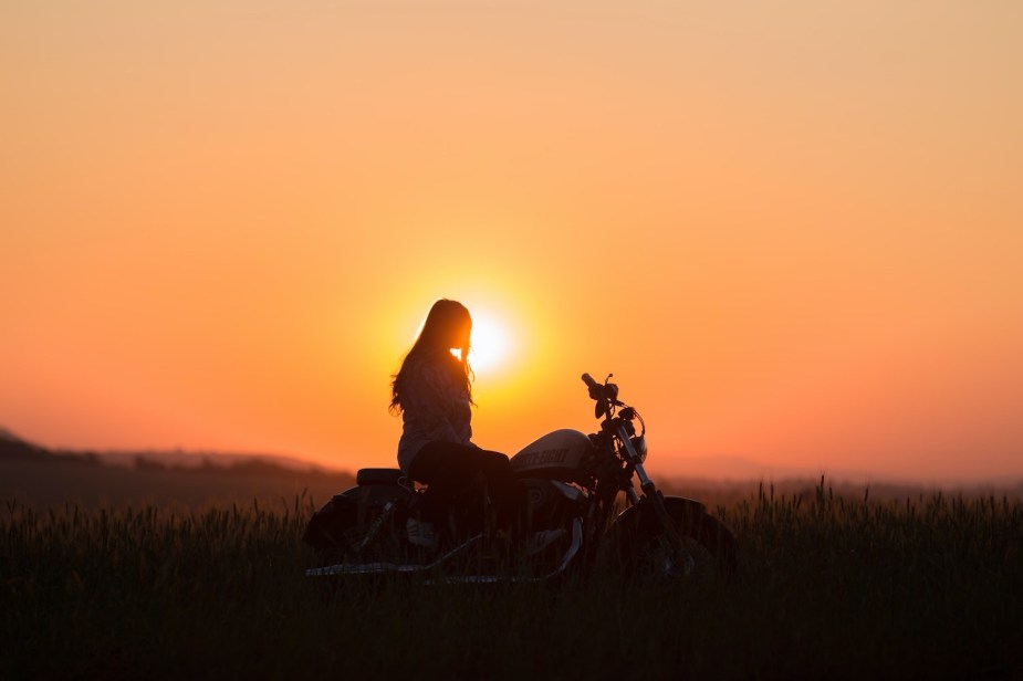 A woman sitting on a motorcycle, outlined against the setting sun, a ridge of mountains visible in the background.