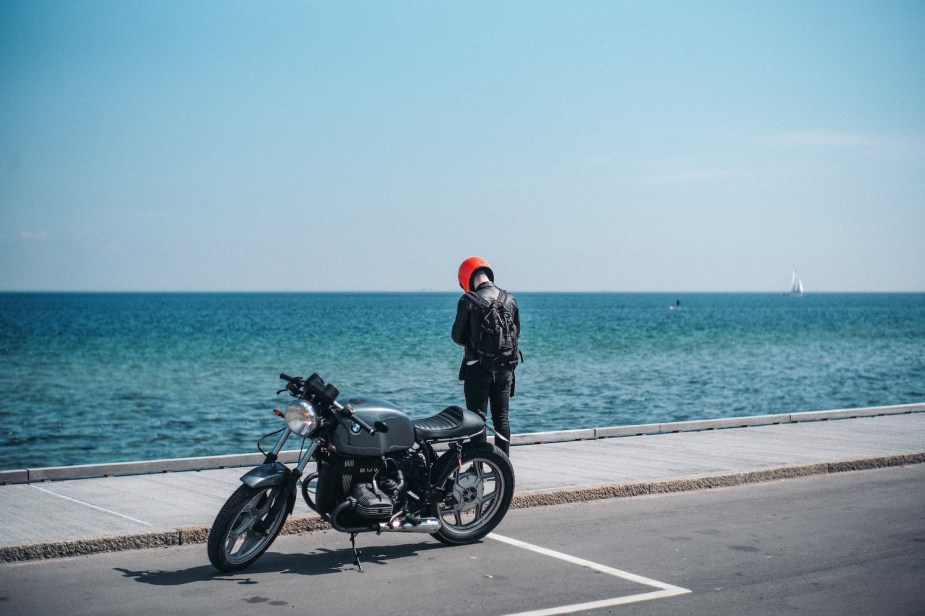 A motorcyclist wearing a backpack stands by their parked bike, looking at the ocean.