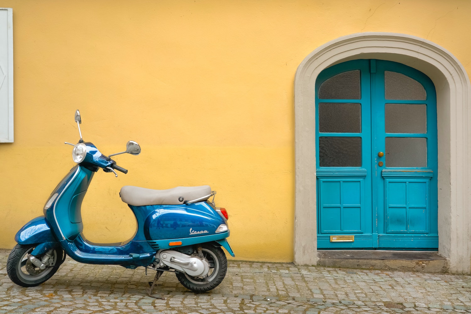 Blue Vespa scooter parked on a cobblestone street with a blue door visible in the background.