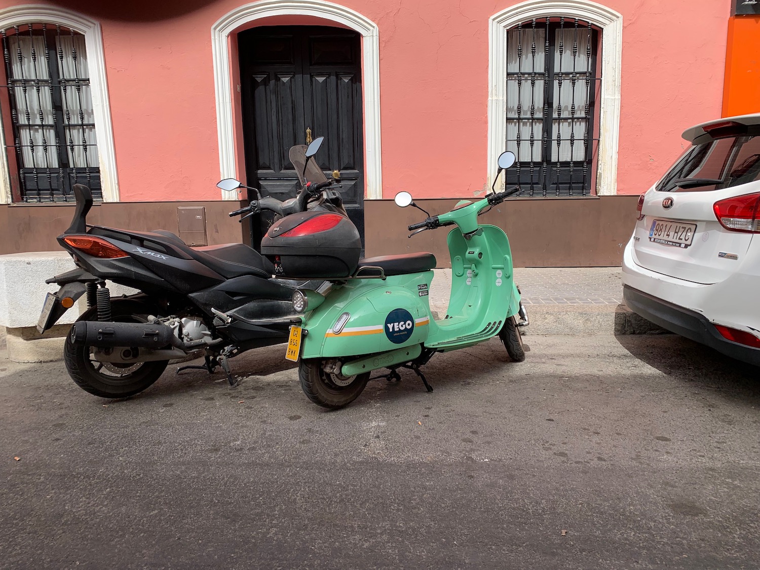 Small, app-based electric rental scooter parked next to a large Moped, a pink building visible in the background.