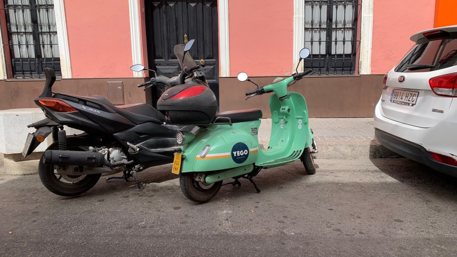 Small, app-based electric rental scooter parked next to a large Moped, a pink building visible in the background.