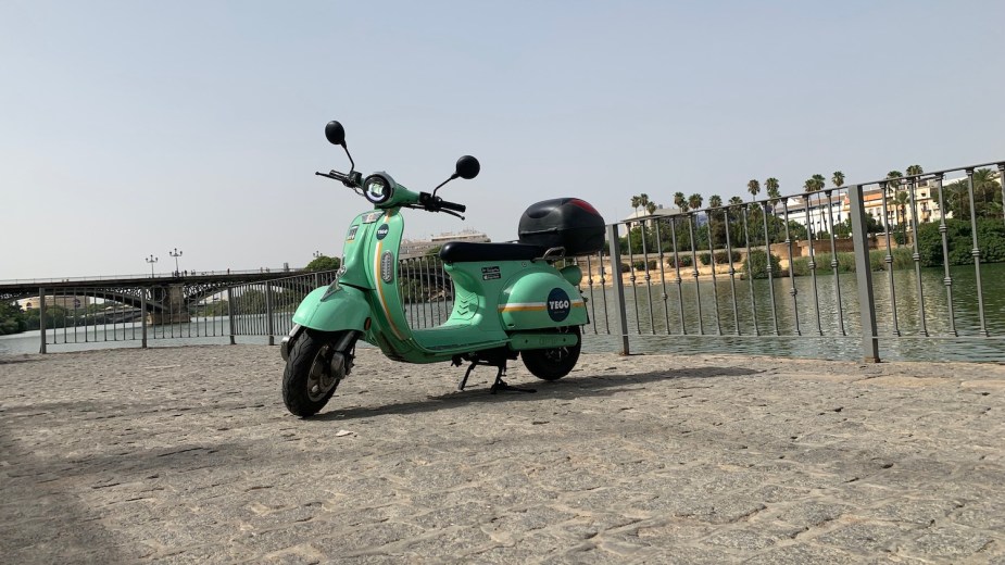 Mint green colored YEGO scooter parked on a cobblestone walkway, a river visible in the background.