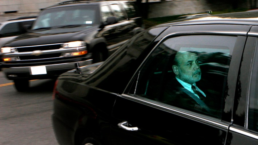 A man in a suit riding in the back seat of a Cadillac car with heavily tinted windows.