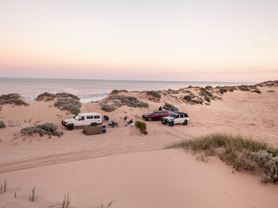 Four SUVs parked on the beach with tents set up around them, dunes and the ocean visible in the background.