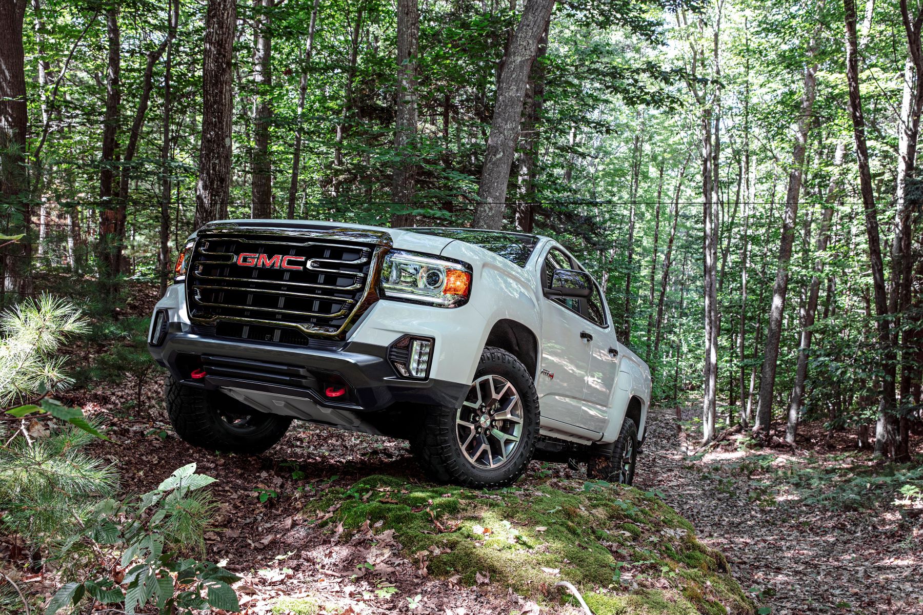 A white 2022 GMC Canyon AT4 midsize truck model (available in diesel) traveling off-road through a forest