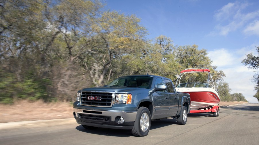 Promo photo of a GMC Sierra full-size hybrid pickup truck towing a boat down a rural road, trees visible in the background.