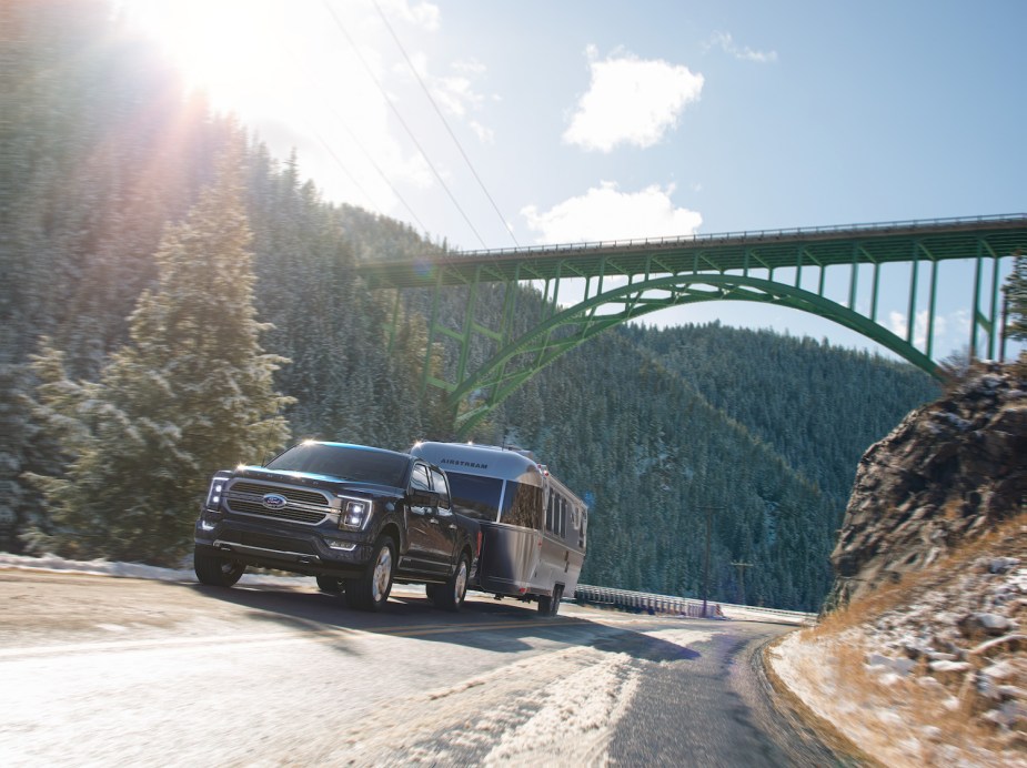 2021 Ford F-150 PowerBoost full-size hybrid pickup truck towing an airstream RV trailer up a mountain road with a bridge visible in the background.