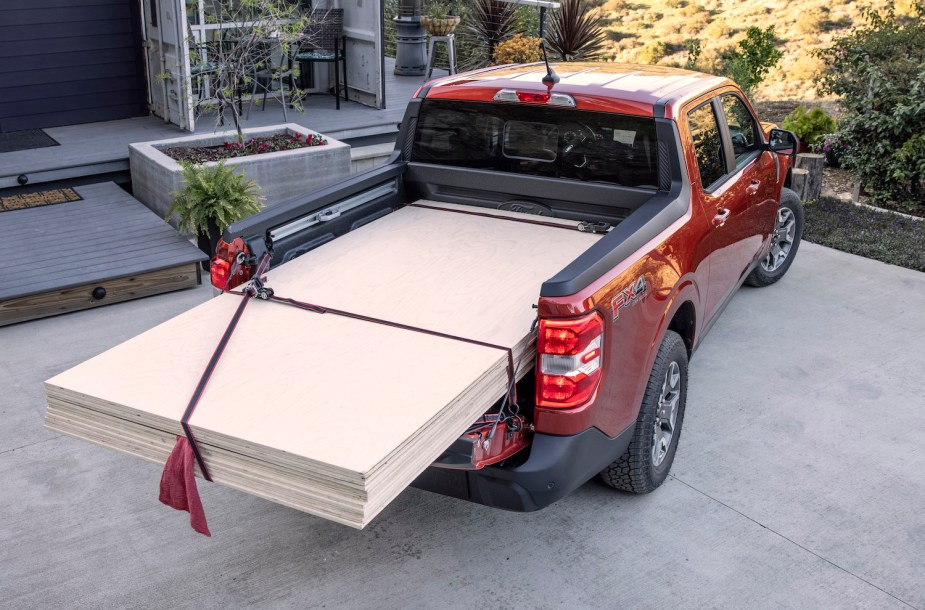 A red Ford Maverick compact truck stands in front of a house with a pile of plywood sheets.