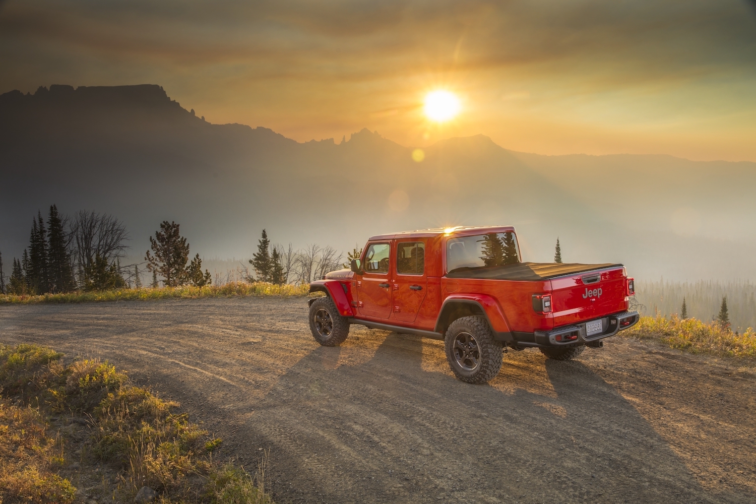 Promotional photo of a red 4WD Jeep Gladiator truck cruising down a dirt road with the sun setting behind a mountain range visible in the background.