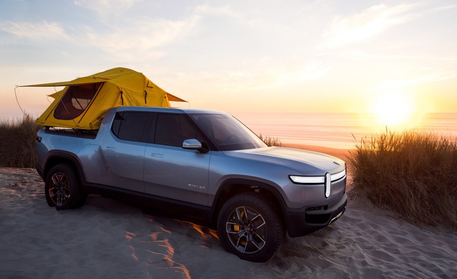 Promo photo of a Rivian electric truck with rooftop tent parked on a sandy beach, the sunset visible in the background.