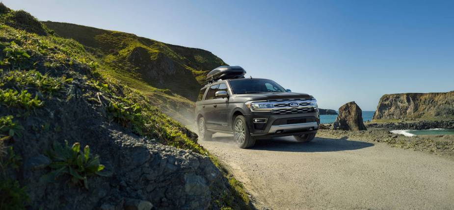 Promo shot of the Ford Expedition SUV  driving along a dirt road with both mountains and the ocean visible in the background.