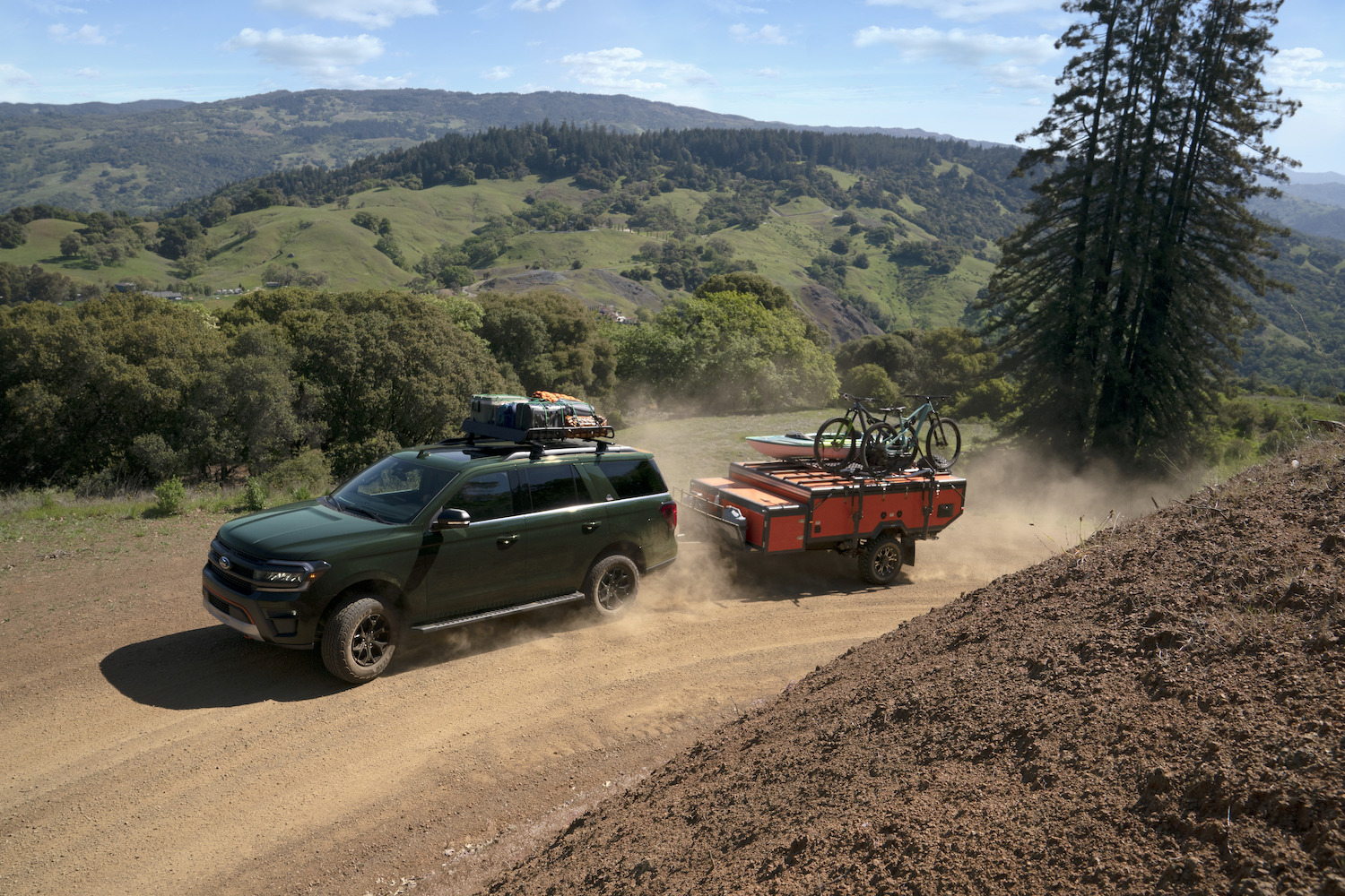 Green Ford Expedition Timberline edition full-size hybrid SUV towing a camping trailer up a dirt road, a mountain pass visible in the background.