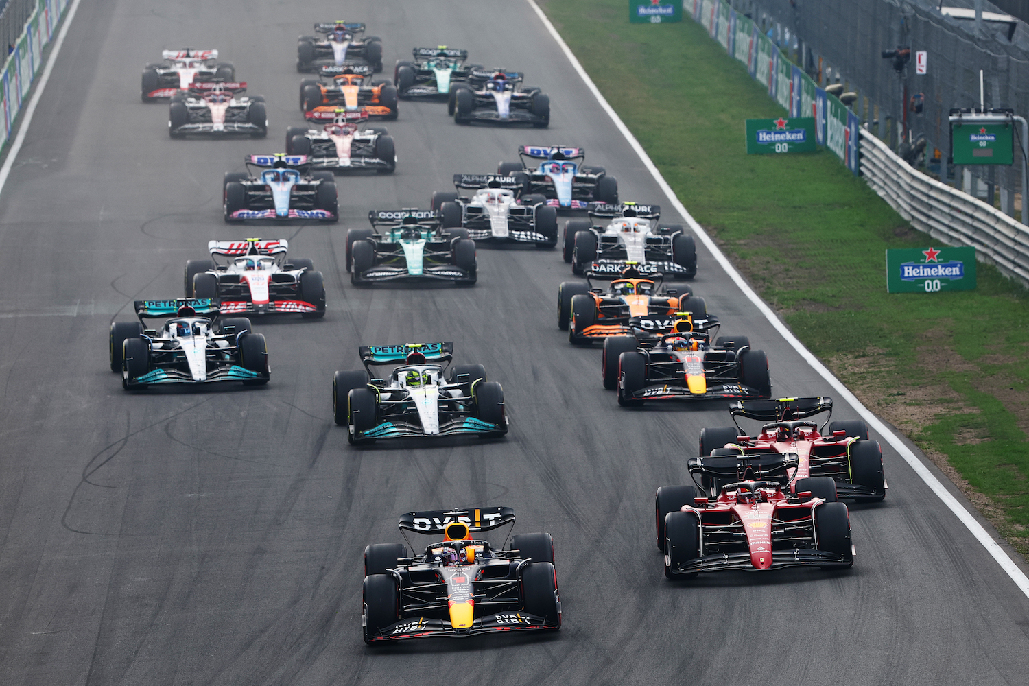 A pack of Formula 1 cars accelerating from the starting line of a race track during a grand prix.