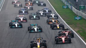 A pack of Formula 1 cars accelerating from the starting line of a race track during a grand prix.