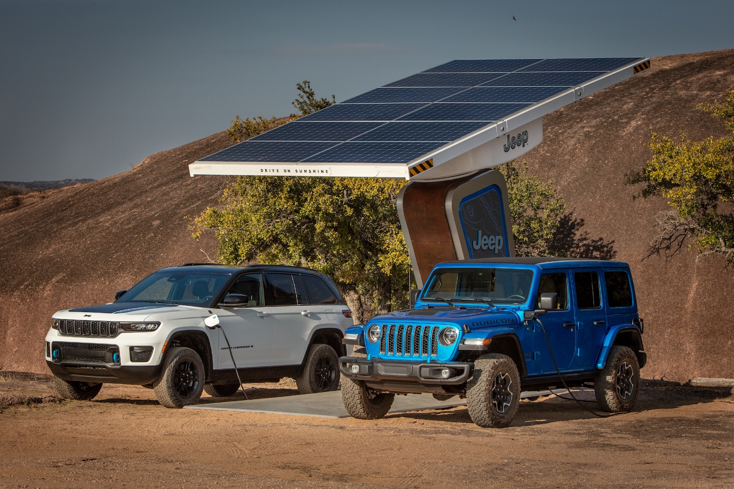Two Jeep hybrid SUVs plugged into a remote solar panel, shrubs and the desert visible behind them.