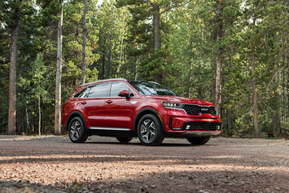 Promo photo of a red Kia Sorento hybrid SUV with a 3rd row of seating parked in a clearing, trees visible in the background.