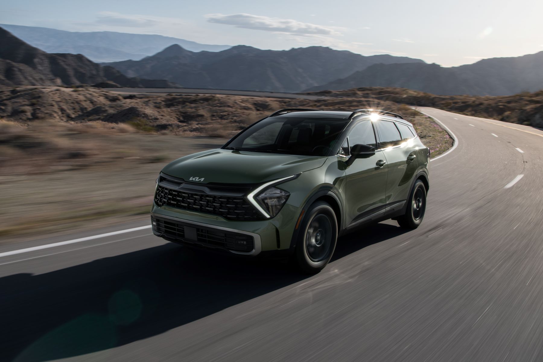 A green 2023 Kia Sportage X-Pro compact SUV driving on a country highway near mountains