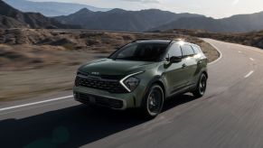 A green 2023 Kia Sportage X-Pro compact SUV driving on a country highway near mountains