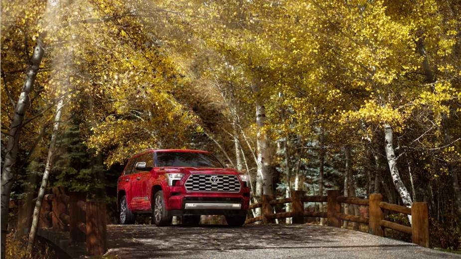 Promo photo of a red Toyota Sequoia hybrid SUV crossing a bridge under a row of birch trees.