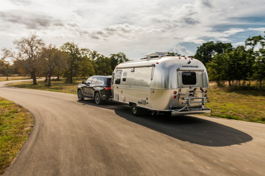 Promo shot of a black Toyota Sequoia hybrid SUV pulling an airstream camper trailer around a bend in a rural road.