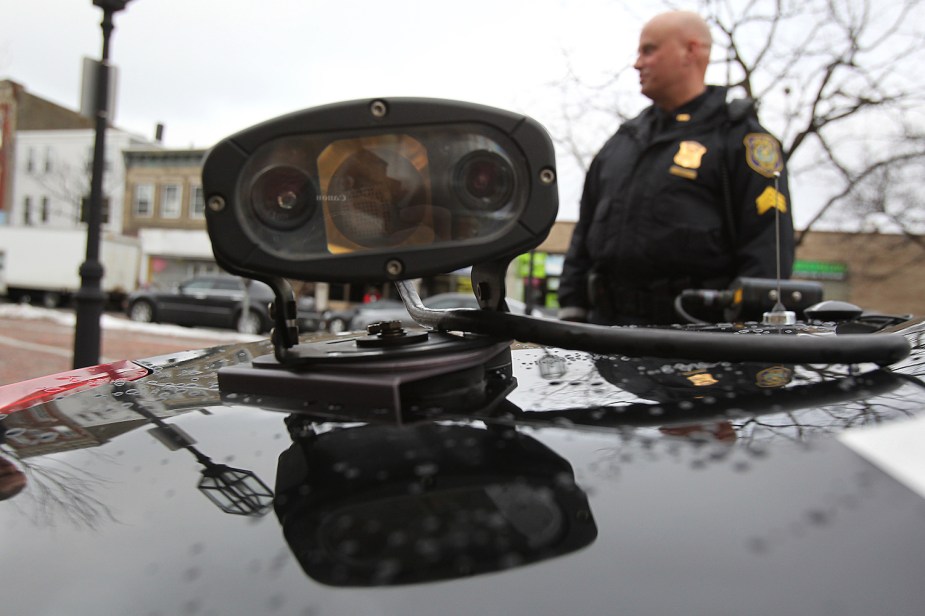 Closeup of an automatic license plate reader systems scanner camera on a car, police officer visible in the background.