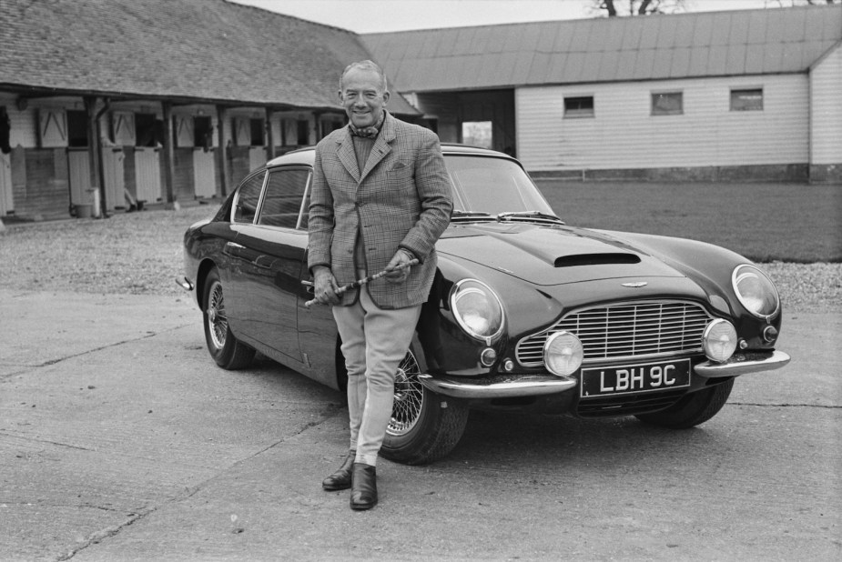 Sir David Brown poses in front of an Aston Martin DB6 grand touring sports car.