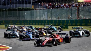 A pack of 2022 Formula 1 race cars rounding a corner during the Belgium Grand Prix, the audience visible in the background.