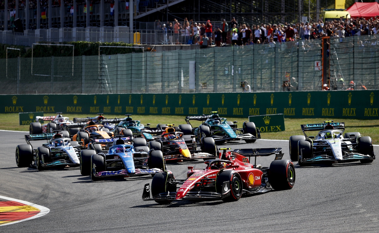 A pack of 2022 Formula 1 race cars rounding a corner during the Belgium Grand Prix, the audience visible in the background.