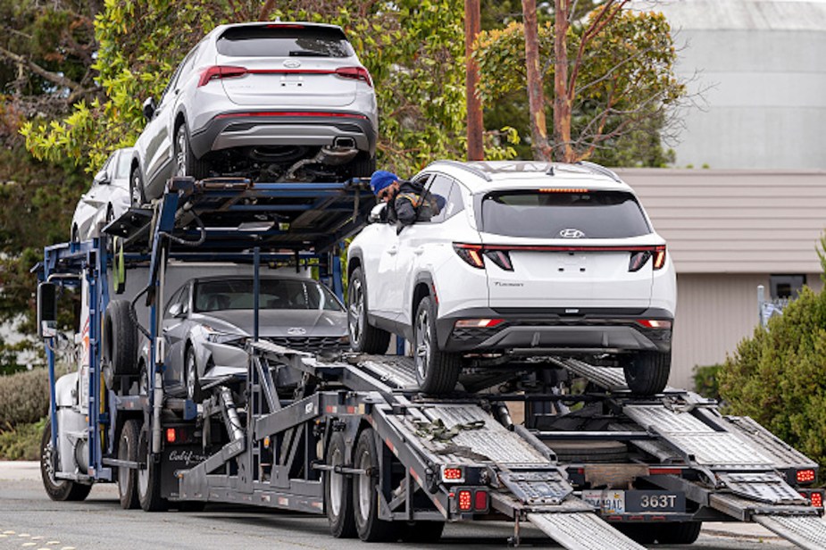 Several cars loaded on a transport trailer, trees and buildings appear in the background.