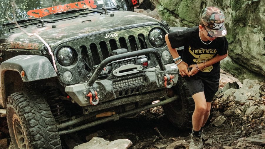 A young man deploys the winch from the front bumper of a 4x4 Jeep SUV, an off-road trail visible in the background.