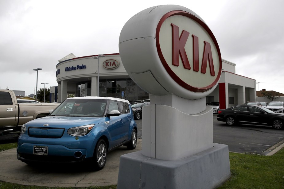 A Kia sign in front of a dealership parking lot, a blue Kia Soul visible behind it.