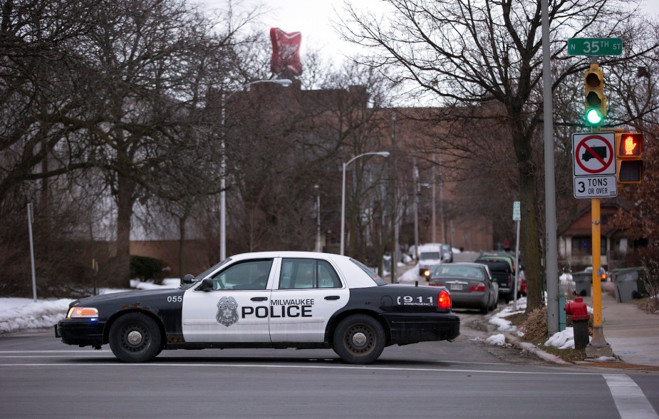 Ford Crown Victory Police-cruiser met de tekst Milwaukee Police rijdt door een kruispunt, gebouwen zichtbaar op de achtergrond.